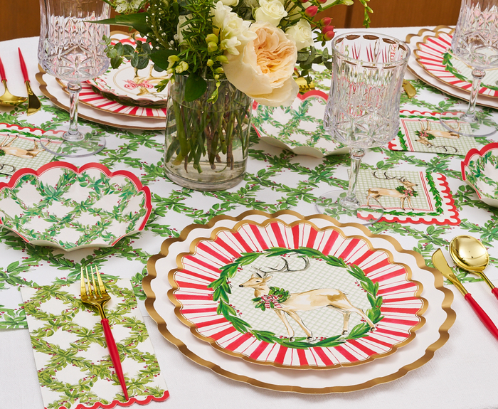 A festive table is set with Sophistiplate Holly Berry Dinner Plates (8pk), red and gold cutlery, patterned napkins, crystal glasses, and a centerpiece of white and yellow flowers atop a green and white tablecloth.