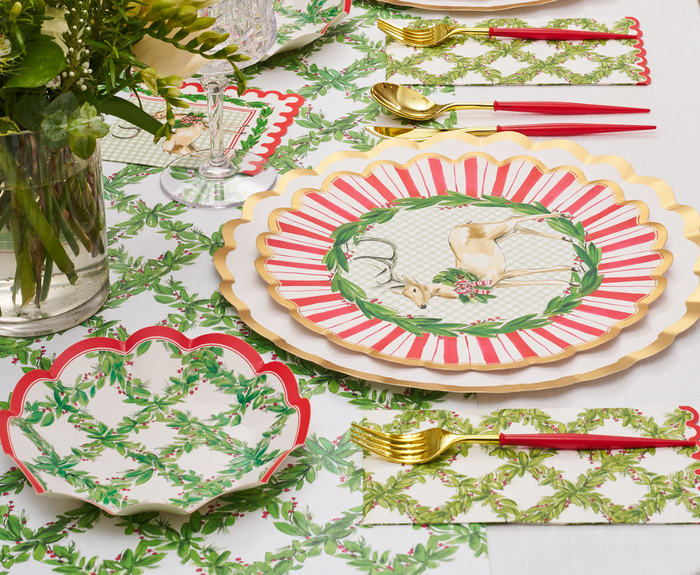A festive table is set with Sophistiplate Holly Berry Dinner Plates (8pk), gold cutlery, red-handled forks, green leafy placemats, and a vase of fresh flowers on the left. Each plate features a charming reindeer illustration in the center.