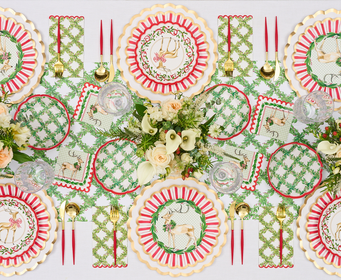 A festive table is set with floral centerpieces, Sophistiplate Holly Berry Dinner Plates, red and green placemats, gold utensils, red candles, clear glassware, white napkins, and a white tablecloth.