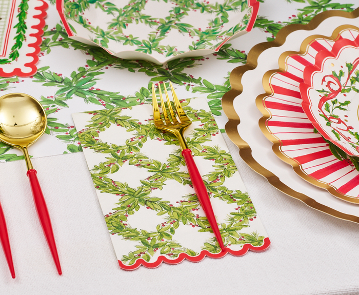 Create a festive table with gold and red cutlery, scalloped plates in red, white, and gold, a bowl of holiday greenery, and Sophistiplate Holly Berry Guest Towels (20pk) on a white tablecloth.