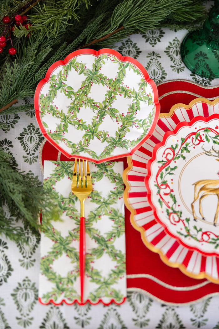 A festive table setting with plates featuring green holly patterns and scalloped red edges, a gold fork with a red handle, Sophistiplate Holly Berry Guest Towels (20pk), and Christmas greenery as decor.