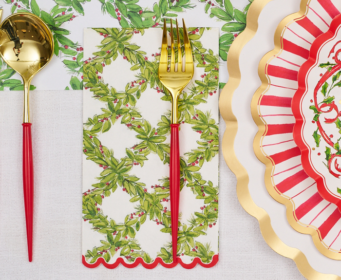 A gold spoon and fork with red handles rest on a Sophistiplate Holly Berry Guest Towel, paired with a red and white striped scalloped plate with a gold border, atop a white and green floral tablecloth.