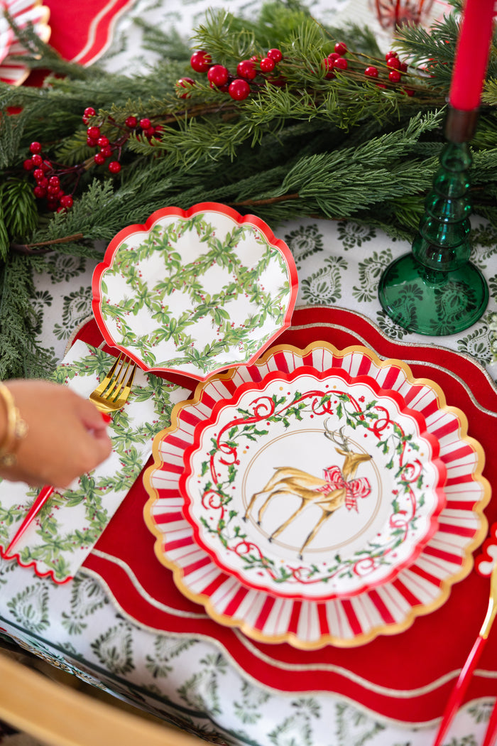 A festive holiday table set with a red placemat, gold utensils, a reindeer plate with red bow, Sophistiplate Holly Berry Guest Towels, a green and white patterned plate, and Christmas greenery with red berry garland centerpiece.