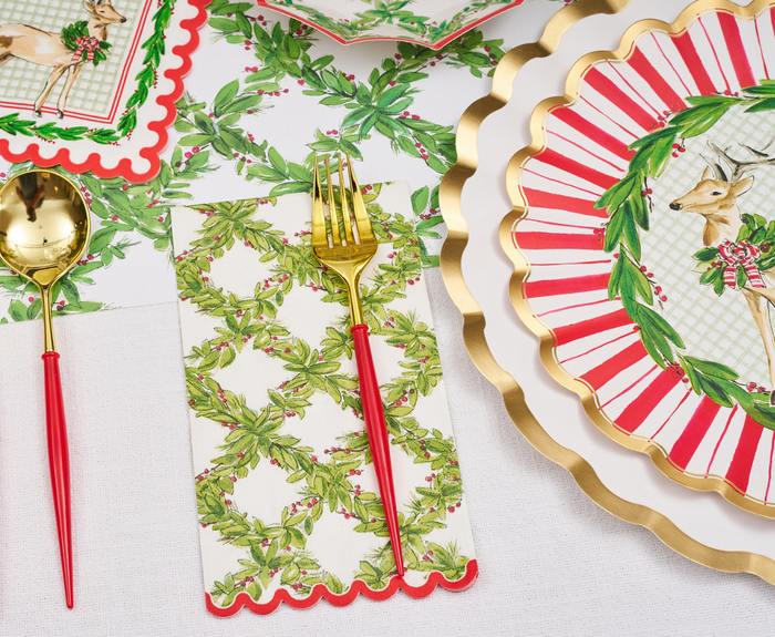 A festive table is set with gold utensils with red handles, holiday-themed plates featuring deer and wreaths, Sophistiplate Holly Berry Guest Towels, Christmas greenery, all arranged on a white tablecloth.