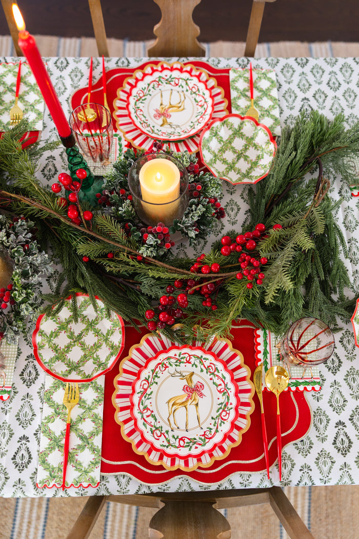 A festive table features Sophistiplate Holly Berry Guest Towels (20pk), Christmas-themed reindeer plates, gold cutlery, a red candle, and a centerpiece of greenery with holly berries on a patterned tablecloth.