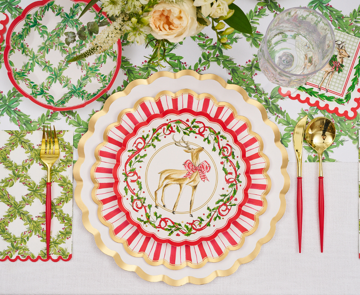 A festive table setting featuring gold and red utensils, Sophistiplate’s Holly Berry Salad Plates adorned with a deer sporting a red bow, and holiday-themed napkins and placemats embellished with green foliage and red accents. The scene is completed by a flower arrangement at the top.