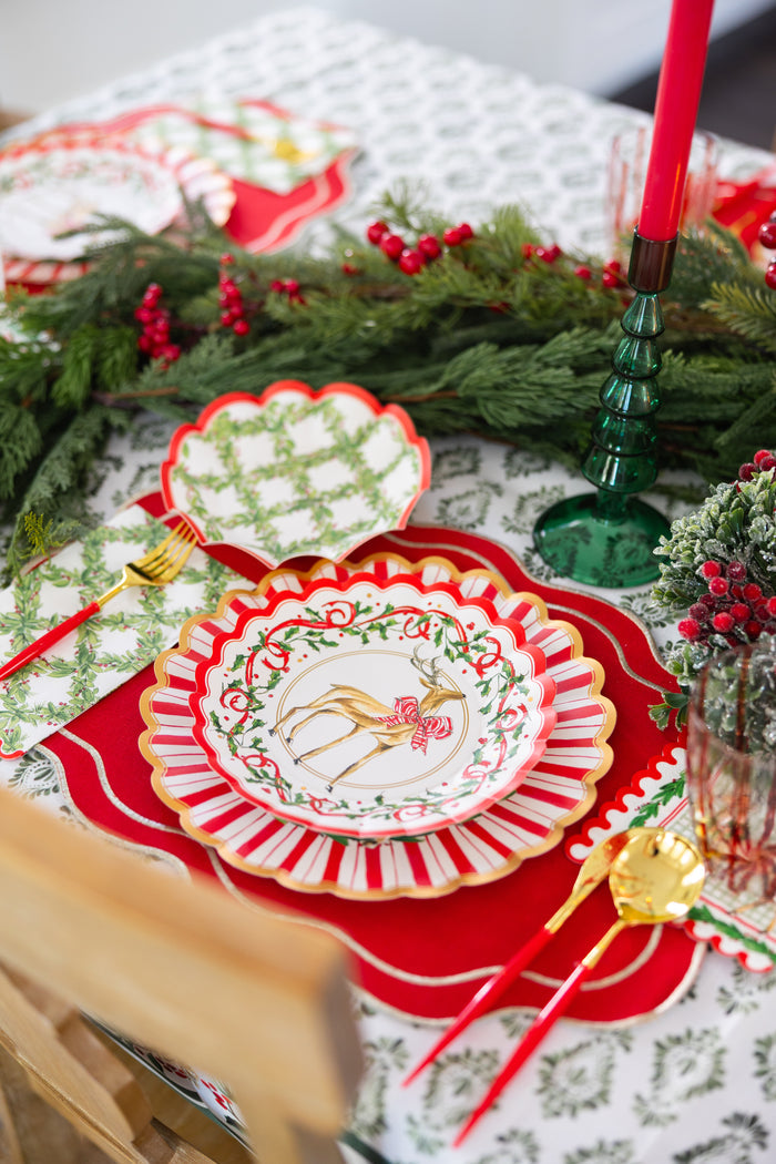 Create a festive holiday table setting with Sophistiplate Holly Berry Salad Plates (8pk), featuring a charming reindeer design, complemented by gold cutlery, red placemats, green glassware, red candles, and garlands adorned with pine, red berries, and lush greenery.
