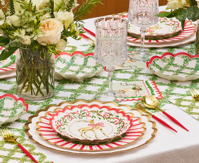 The table is festively set with Sophistiplate Holly Berry Salad Plates (8pk), gold utensils, crystal glasses, and a white tablecloth featuring green patterns. A bouquet of white and cream flowers serves as the centerpiece.