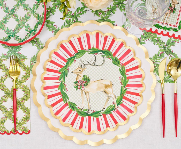 The Sophistiplate Holly Berry Table Setting features gold utensils, reindeer and wreath designer plates, holly napkins, a clear glass, and a white holiday-themed tablecloth for a festive Christmas tablescape.