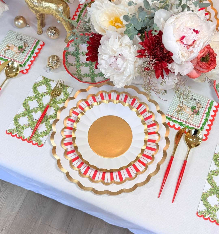 Create a festive holiday table with Sophistiplate’s Holly Berry Table Setting, featuring gold and white designer plates, gold cutlery with red handles, napkins with greenery and reindeer, and a floral centerpiece on a crisp white tablecloth.
