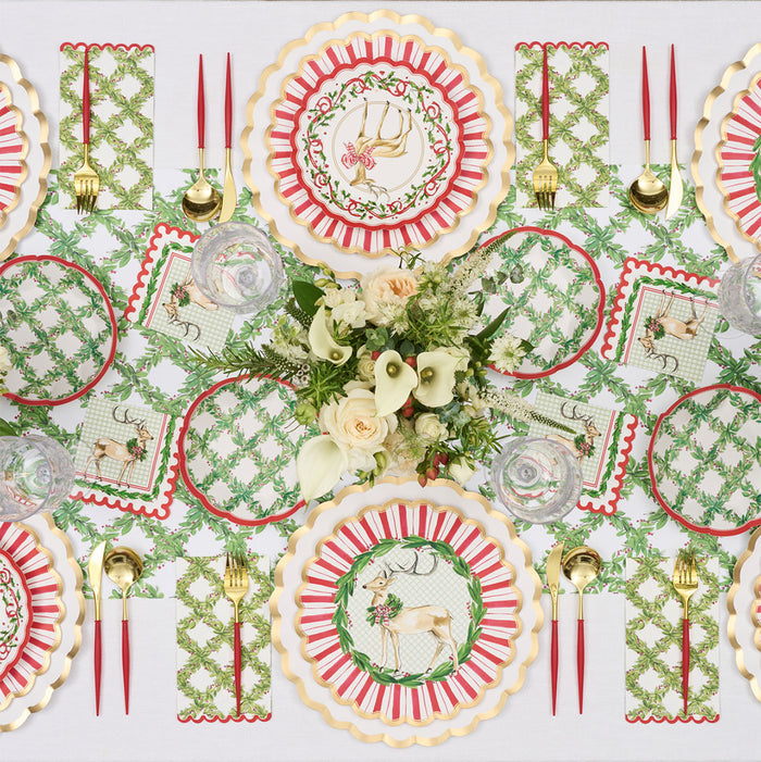 A festive Christmas table features red, gold, and green reindeer and monogram plates, gold utensils, crystal glasses, and a centerpiece of white calla lilies and roses on the Rosanne Beck Holly Berry Trellis Table Runner (20x12").
