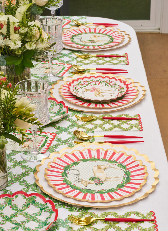 A festive table is set with gold-rimmed plates, red and white stripes, floral and bird designs, gold cutlery with red handles, crystal glasses, and the Rosanne Beck Holly Berry Trellis Table Runner (20x12") atop a white cloth with green leafy accents.