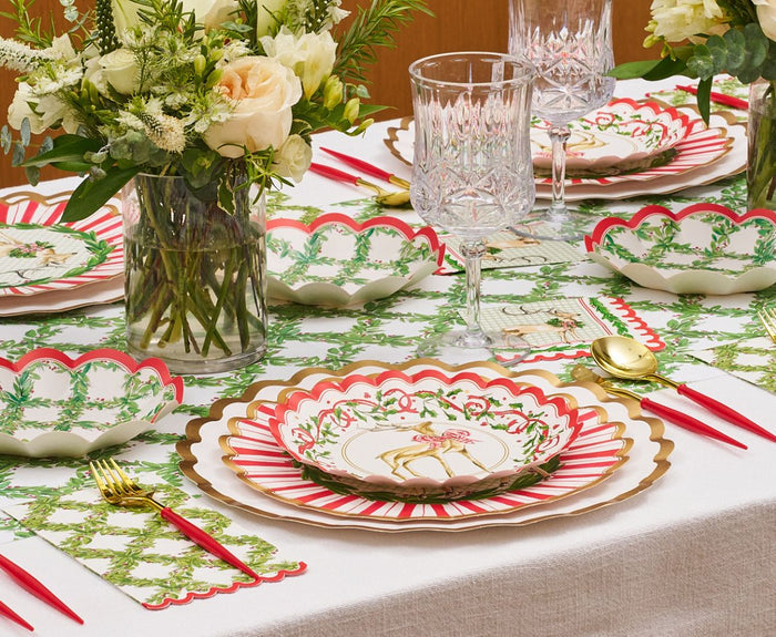 A festive Christmas table features the Rosanne Beck Holly Berry Trellis Table Runner (20x12"), floral arrangements, scalloped red, white, and gold plates, green placemats, gold and red cutlery, and crystal glasses on a white cloth.