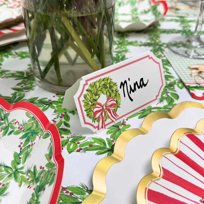 A festive table is set with a Rosanne Beck Holly Wreath Place Card labeled Nina, surrounded by holly plates with gold scalloped edges, and a vase of green stems in the background.