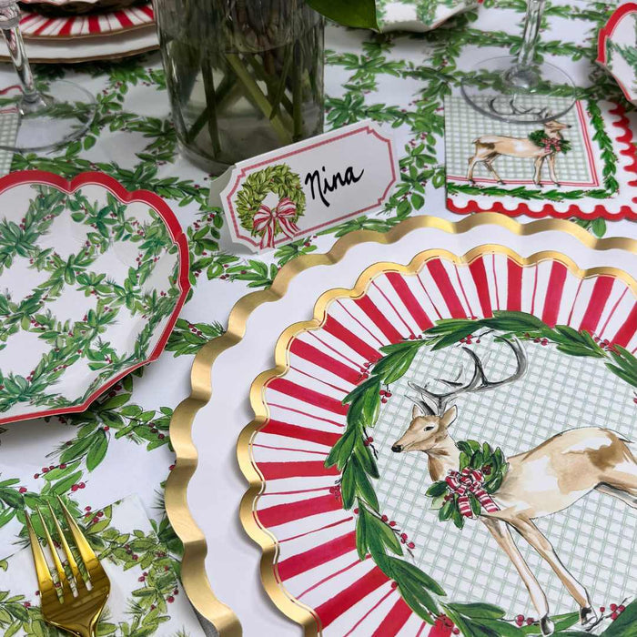 A festive table features Rosanne Beck plates with a deer and holly wreath, red and white striped borders, a green and white tablecloth, gold cutlery, and Rosanne Beck Holly Wreath Place Cards labeled "Nina.