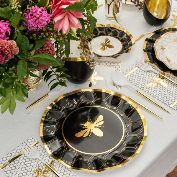 A table set featuring Sophistiplate Honeybee Salad Plates (8pk) showcases charming black and gold ruffled edge paper plates with a honeybee motif, complemented by gold cutlery, bee-printed napkins, and a vibrant floral centerpiece of pink and red blooms. The decor is enhanced with honeycomb patterns and elegant gold accents.