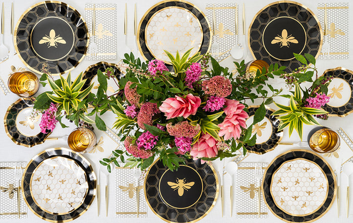 A table is set with Sophistiplate Honeybee Salad Plates, featuring a black and gold honeybee design and charming ruffled edges, complemented by gold cutlery and yellow glasses. The centerpiece displays pink and purple flowers with green leaves, while the entire arrangement is neat and symmetrical.