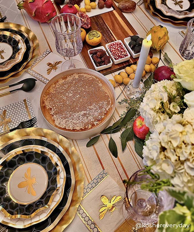 A festive table set with gold and black paper plates—some featuring a subtle honeybee design—alongside a bowl of honeycomb, fruits, flowers, and lit candles, arranged for a celebration. Pomegranates, figs, and a challah cover are visible among the party tableware decorations.