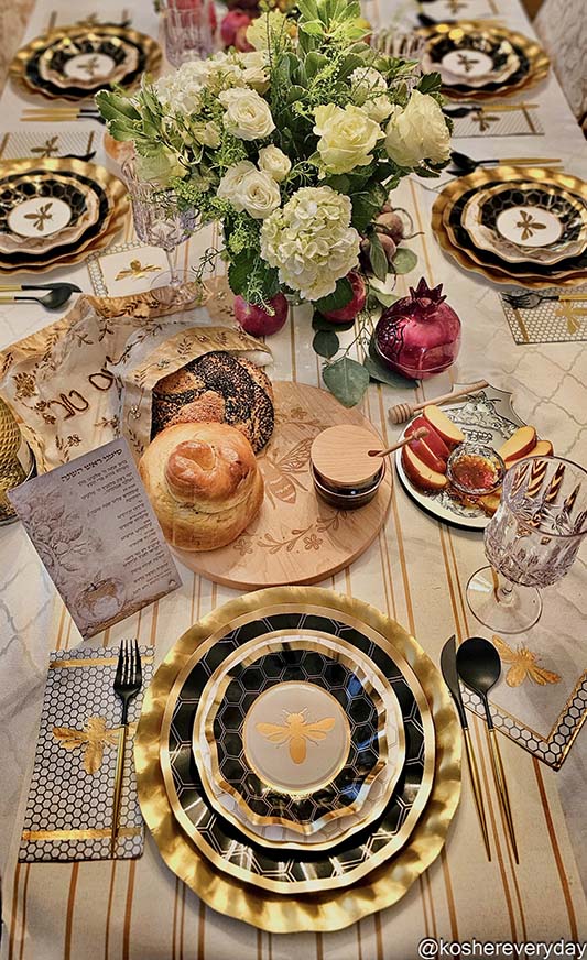 A festive table is set with Sophistiplate Honeybee Dinner Plates (8pk), featuring ruffled edges and gold and black accents, accompanied by challah bread, honey, apples, pomegranate, wine glasses, and a floral centerpiece.