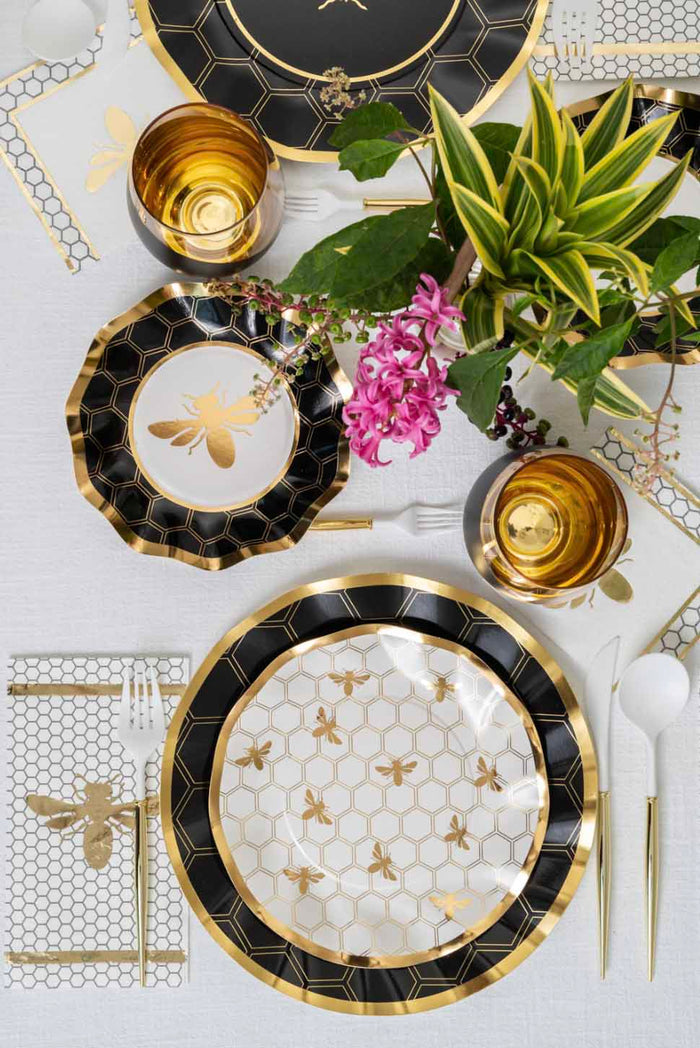 Table setting with black, white, and gold bee-themed plates, gold cups, white cutlery, Sophistiplate Honeybee Guest Towels (20 pack) featuring an elegant honeybee design, and a potted plant with pink flowers on a white tablecloth.