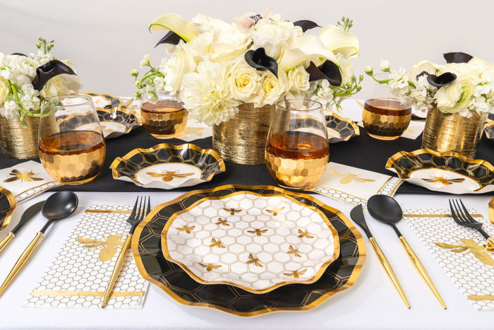 Experience an elegant table setting featuring Sophistiplate’s Honeybee Salad Plates/8pk, showcasing a chic black, gold, and white honeybee design with ruffled edges. The sophisticated ensemble is completed with gold flatware, black napkins, honeycomb-patterned placemats, amber glasses, and white floral centerpieces arranged in gold vases—perfect for a formal event.