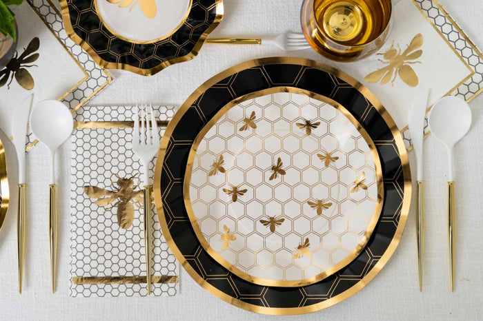 Elegant table setting featuring Sophistiplate Honeybee Salad Plates (8pk) with ruffled edges, gold utensils, a clear glass filled with amber liquid, white cutlery, and a bee-patterned napkin on a crisp white tablecloth.