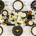 The Honeybee Table Setting by Sophistiplate LLC showcases bee-designed paper plates in black, gold, and white, gold utensils, amber glasses, and white florals symmetrically arranged on a black table runner.