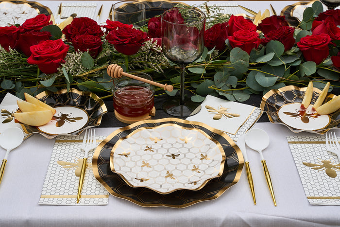 Elegant table setting with bee-themed party tableware, including honeybee design paper plates, gold utensils, red roses, a jar of honey with a dipper, and sliced apples on small plates. The décor features black, white, and gold accents.