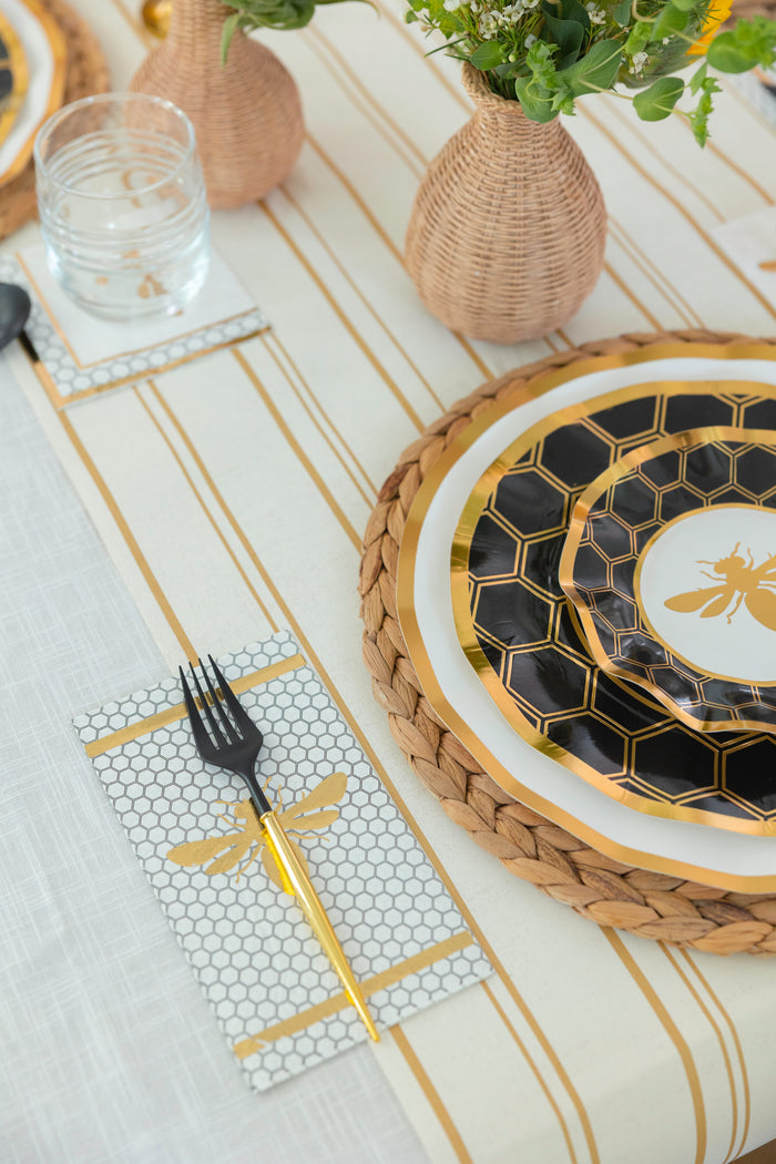 The Honeybee Table Setting by Sophistiplate LLC features gold and black heavy-duty paper plates with a bee design, a gold fork on a honeycomb-patterned napkin, woven vases with flowers, and a striped tablecloth.