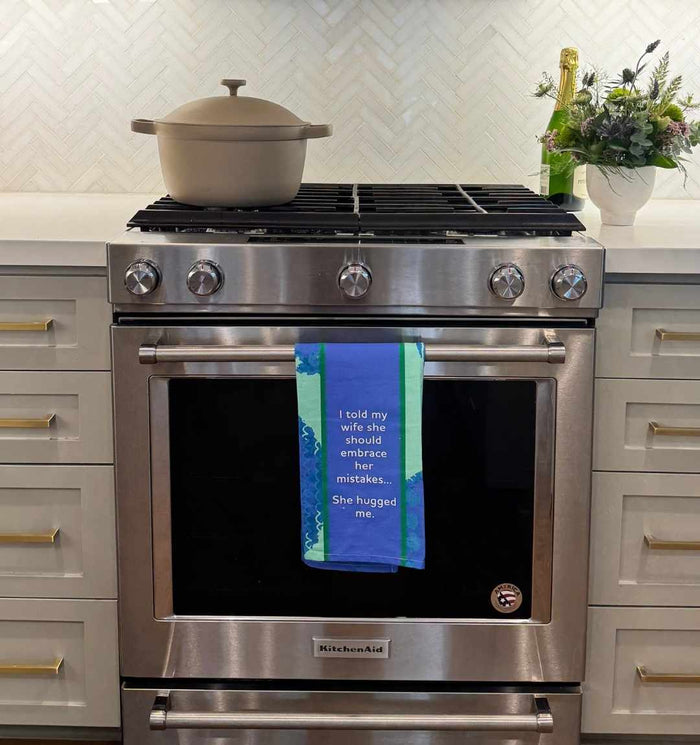 A kitchen with a stainless steel oven features the Drinks On Me "Hugged Me Dish Towel," reading, “I told my wife she should embrace her mistakes... She hugged me.” A beige Dutch oven sits on the stove; flowers and a bottle are on the counter.