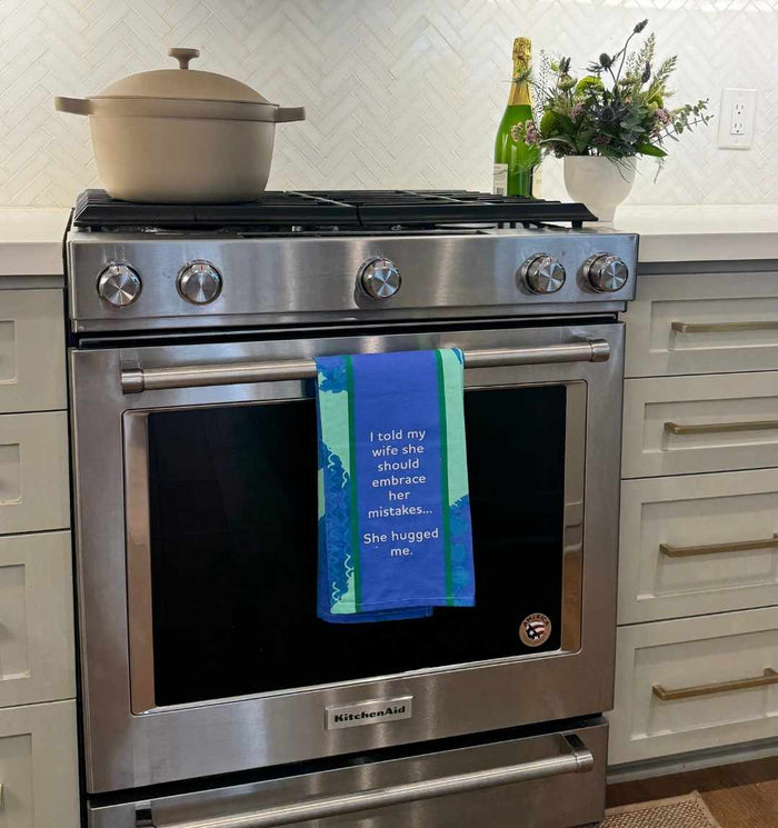 A kitchen stove features a beige pot, green bottle, and flower arrangement on the counter. Hanging from the oven is the Drinks On Me "Hugged Me Dish Towel," reading: “I told my wife she should embrace her mistakes… She hugged me.”.