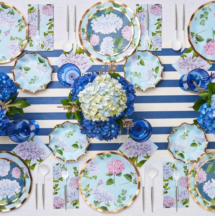 The Hydrangea Table Setting by Sophistiplate features a blue and white striped tablecloth, floral designer plates, blue glasses, gold-rimmed dishes, white cutlery, matching floral napkins, and a blue and white hydrangea centerpiece.