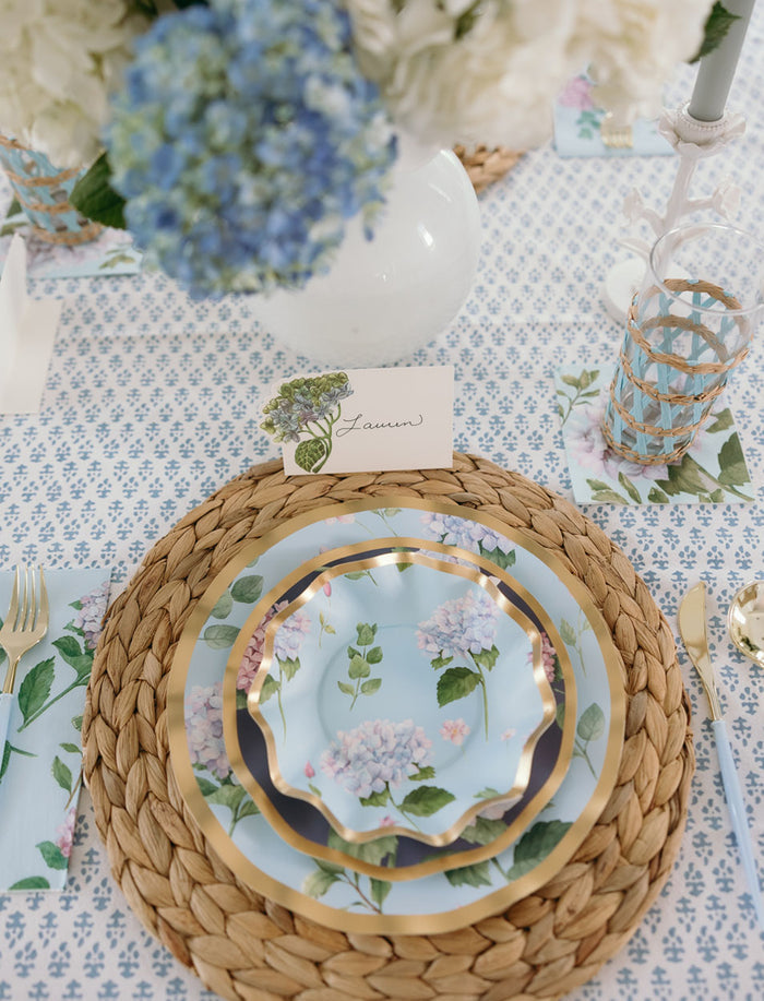 The Hydrangea Table Setting by Sophistiplate features gold-rimmed designer disposable plates on a woven charger, gold utensils, a floral napkin, a blue-patterned water glass, a “Lauren” name card, and hydrangeas.
