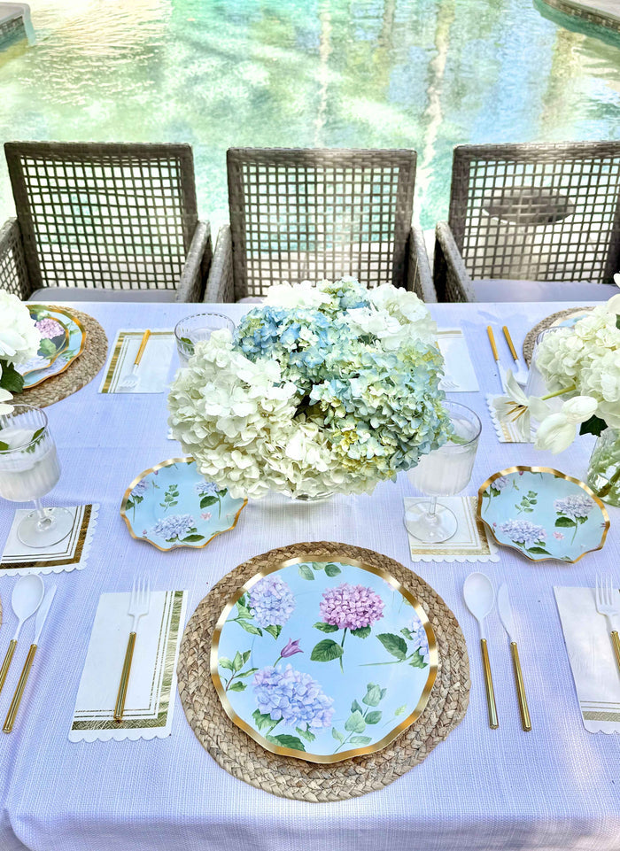 The Hydrangea Table Setting by Sophistiplate features designer disposable plates, gold cutlery, woven placemats, white napkins, and a white and blue hydrangea centerpiece, elegantly displayed outdoors by the pool.