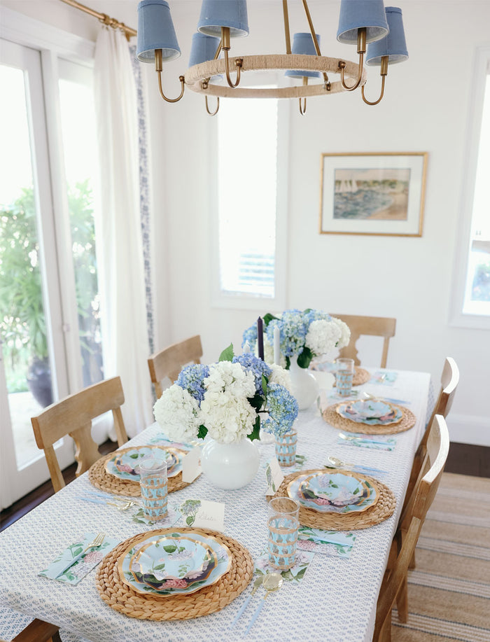 The Hydrangea Table Setting by Sophistiplate features a dining table for four with designer disposable plates, blue and white glassware, and white vases of hydrangeas. Sunlight from nearby windows creates a bright, airy ambiance.