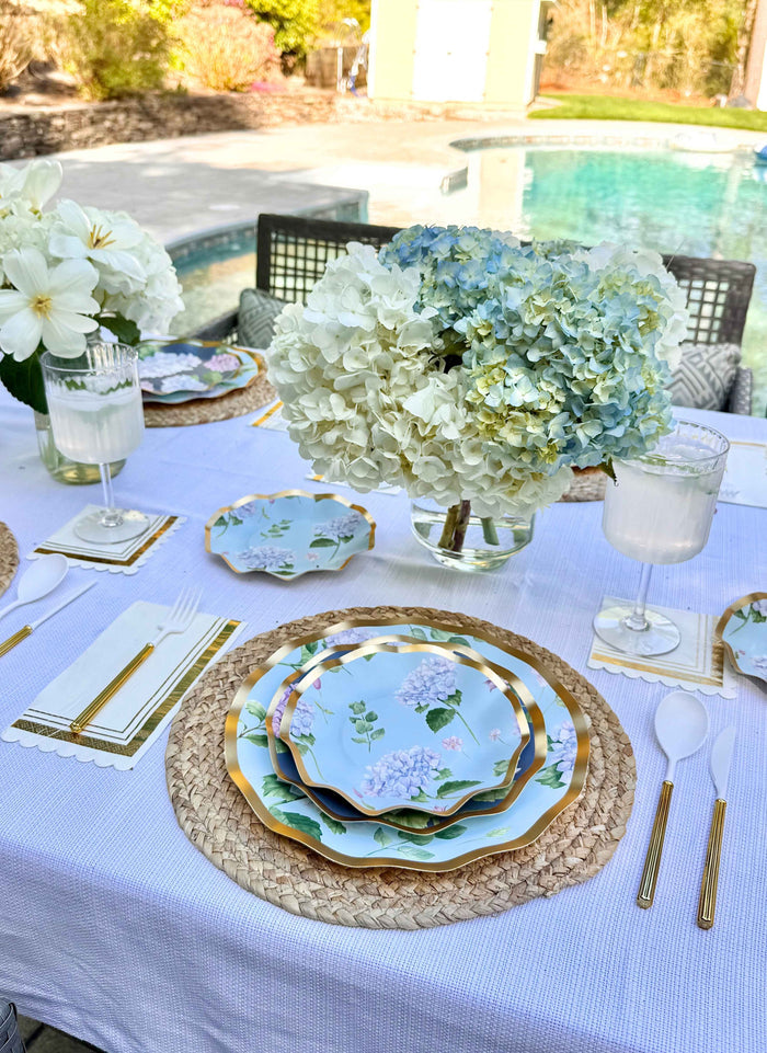 The Hydrangea Table Setting by Sophistiplate features floral designer disposable plates, gold cutlery, woven placemats, white napkins, water glasses, and lush hydrangea bouquets—perfect for elegant outdoor dining by the pool and garden.