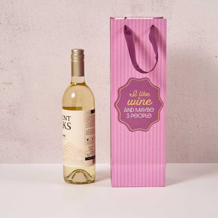 A bottle of white wine sits beside the Drinks On Me "I Like Wine" wine bag with purple handles and the quote "I like wine and maybe 3 people." Perfect as a hostess gift, both rest on a white surface against a light background.