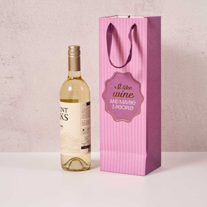 A white wine bottle sits beside the Drinks On Me "I Like Wine" wine bag, featuring handles and the phrase "I like wine and maybe 3 people," making it a great hostess gift against a light background.