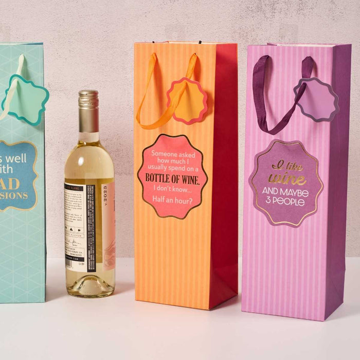 The Drinks On Me "I Like Wine" wine bags come in four colors with ribbon handles and funny sayings, shown upright beside a white wine bottle—perfect hostess gifts delivered in style.