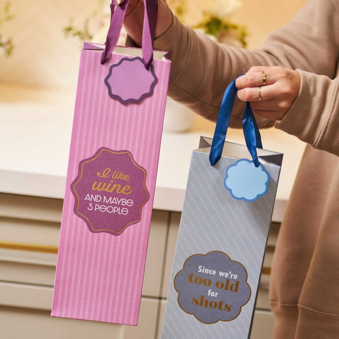 A person holds two Drinks On Me wine bags: one pink "I Like Wine" bag and a gray "Since We're Too Old for Shots" bag, both with ribbon handles and funny labels—perfect hostess gifts.