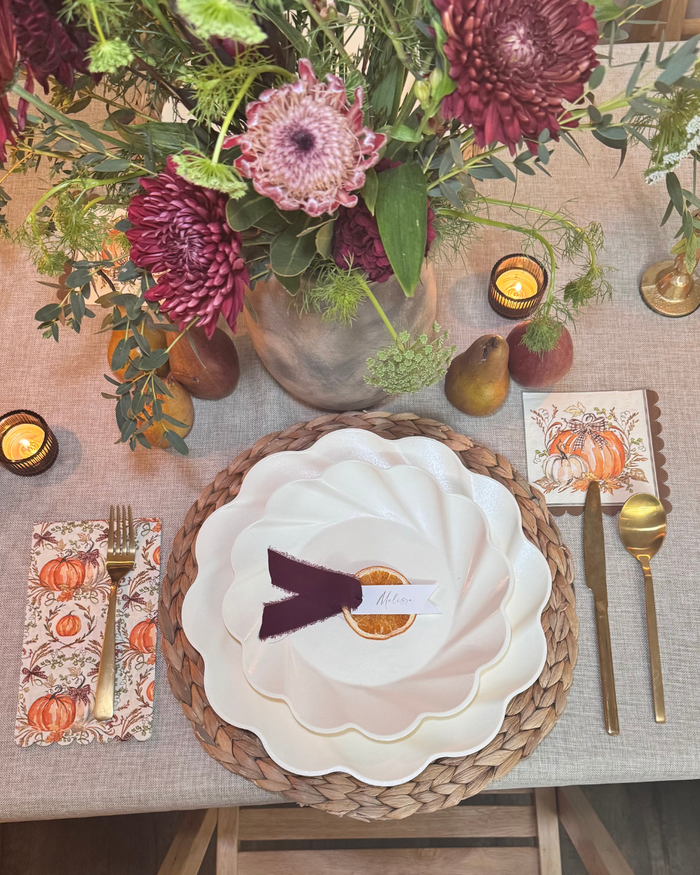 Create a welcoming autumn atmosphere with the Ivory Harvest Table Setting by Sophistiplate. This fall-inspired place setting features gold cutlery, pumpkin-themed napkins, layered white plates atop a woven mat, a name card, pears, candles, and a lush floral arrangement of chrysanthemums and greenery. Elevate your Thanksgiving celebration with Thanksgiving tableware like eco-friendly disposable or compostable plates from Sophistiplate—allowing you to entertain in style while embracing sustainability.