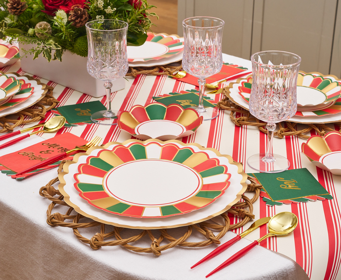 A festive table set with Sophistiplate Joyful Christmas Dinner Plates/8pk, gold utensils with red handles, crystal glasses, a striped red-and-white tablecloth, green name cards, and a floral centerpiece.