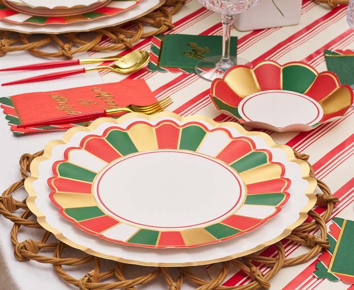 A festive table is set with Joyful Christmas Dinner Plates by Sophistiplate, red, green, white, and gold bowls, gold cutlery, woven placemats, a striped tablecloth, and holiday napkins—perfect for your holiday feast.