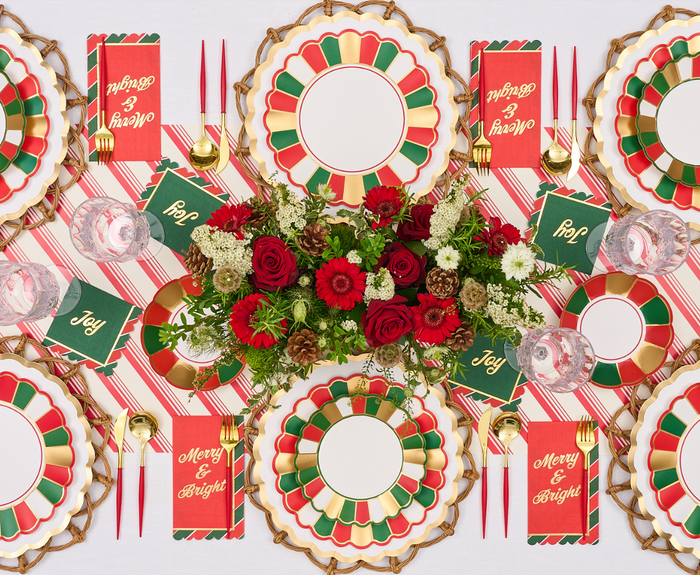 Create a festive holiday table with Sophistiplate Joyful Christmas Salad Plates (8pk), gold flatware, striped napkins, "Joy" and "Merry & Bright" cards, plus a centerpiece of red and white flowers.