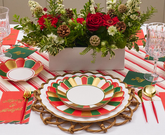 A festive holiday table features Sophistiplate’s Joyful Christmas Salad Plates (8pk) in red, green, and gold, paired with gold cutlery, crystal glasses, a centerpiece of red roses, pinecones, white flowers, plus striped placemats and festive napkins.