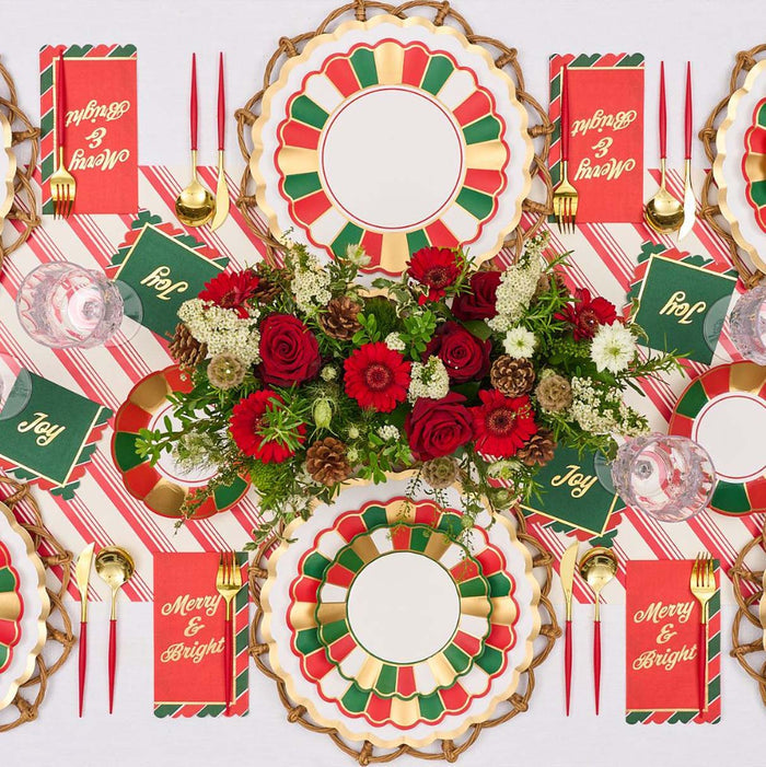 The Joyful Christmas Table Setting by Sophistiplate includes red, green, and gold disposable plates, Merry & Bright and Joy napkins, gold cutlery, red candles, and a festive centerpiece—perfect for holiday gatherings.