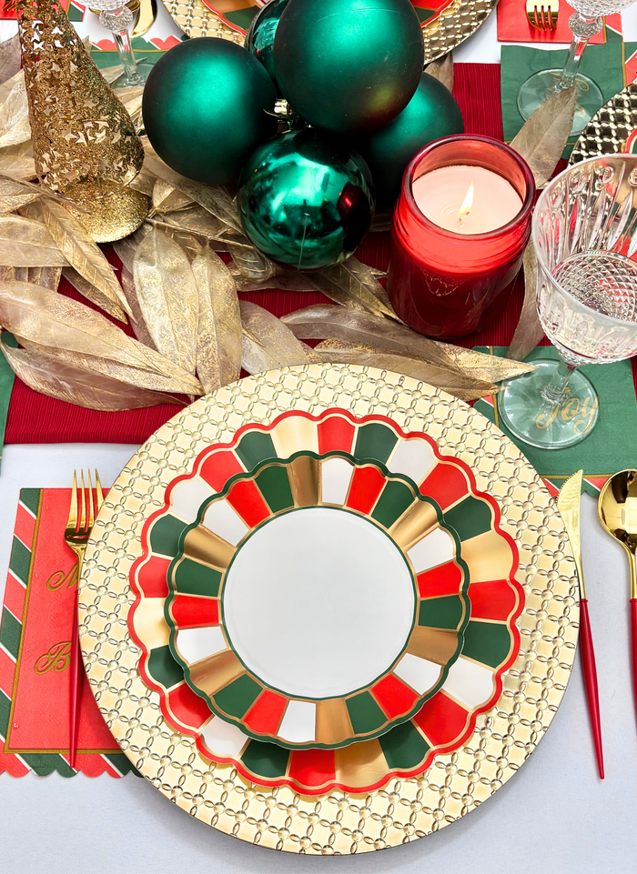 The Sophistiplate Joyful Christmas Table Setting includes a gold-patterned charger, red, green, and gold-rimmed plate, gold flatware, red candle, green ornaments, gold ribbon, and Christmas-themed napkins for a festive tablescape.