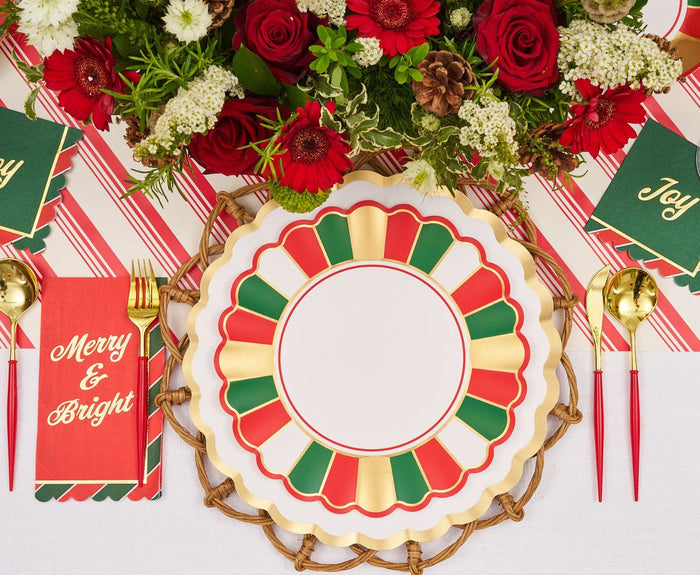 The Joyful Christmas Table Setting by Sophistiplate includes a red, green, and gold plate, gold utensils, “Merry & Bright” napkin, striped tablecloth, centerpiece of red and white flowers, and Joy cards for festive holiday gatherings.