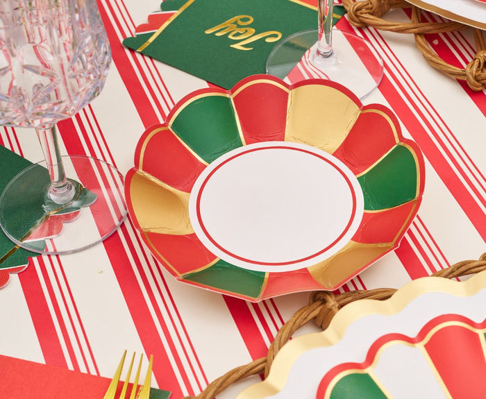 The Sophistiplate Joyful Christmas Table Setting includes a red, green, and gold scalloped disposable plate, clear textured glass, gold utensils, green “Joy” napkins, and a red/white striped tablecloth—perfect festive party supplies.