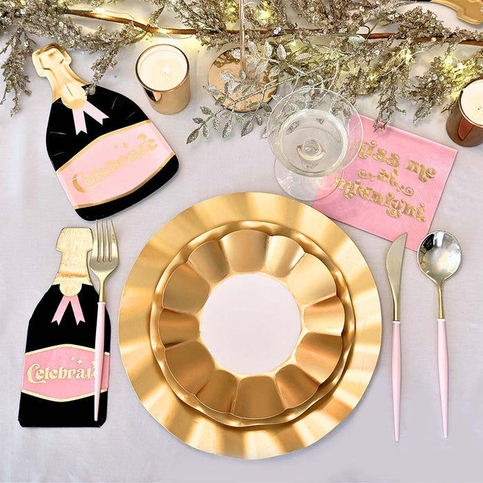 The Sophistiplate Kiss Me at Midnight Table Set includes designer disposable plates, pink and gold cutlery, "Kiss me at midnight" napkins, champagne bottle decorations, candles, and gold foliage on a white tablecloth.